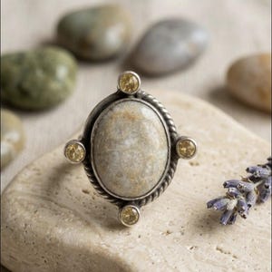 May include: A silver ring featuring a large, oval-shaped, beige fossilized coral cabochon at the center. The setting is accented with four small, round, yellow faceted stones placed at the top, bottom, and sides, surrounded by a twisted rope border.