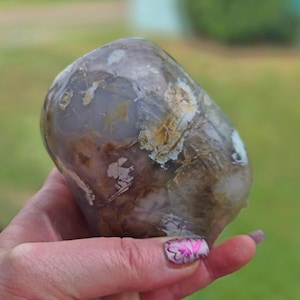May include: A polished, rounded stone featuring a translucent, muted lavender and grey base color. The surface displays intricate, organic inclusions in shades of earthy brown, cream, and white, creating a marbled, multi-layered appearance.
