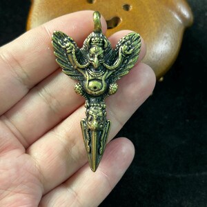May include: A detailed brass pendant featuring a bird-like deity with outstretched wings and a crown. The pendant has a dagger-like bottom and a loop at the top for a chain. The metal has an aged, antique finish.