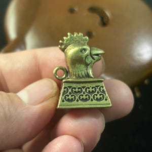Peut inclure: Un petit charme en laiton antique en forme de tête de coq. La tête de coq présente des détails, notamment une crête et un bec. Le charme est monté sur une base décorative avec un motif orné. Une petite boucle est attachée sur le côté de la tête du coq.