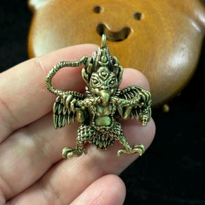 Op de afbeelding: Een gedetailleerde messing hanger met een mythisch vogelachtig wezen met uitgestrekte vleugels en ingewikkelde details. De hanger heeft een lus aan de bovenkant om op te hangen en wordt in een hand gehouden. De achtergrond is wazig.