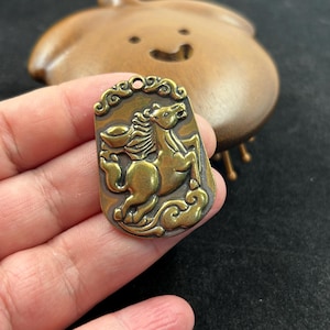 May include: A rectangular, antique brass pendant featuring a detailed relief of a rearing horse. The horse is surrounded by decorative clouds and swirls. The pendant has a small hole at the top for hanging. The background is blurred.