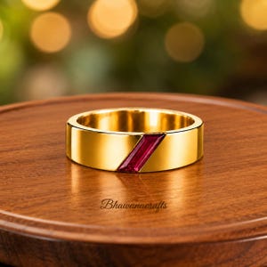 Peut inclure: Une bague anneau en or avec une pierre précieuse rouge taille baguette, sertie en diagonale dans la surface métallique. La bague présente une finition lisse et polie avec un profil plat. Le texte Bhawanacrafts est visible sous la bague.