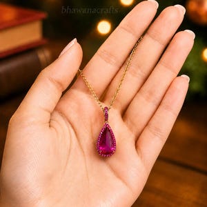 May include: A gold necklace featuring a magenta teardrop pendant, encircled by smaller magenta stones. The necklace is displayed in an open hand. The background includes the text "bhawanacrafts".