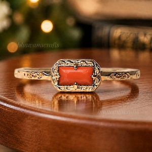 May include: A gold-colored ring with an orange rectangular stone. The ring has an ornate design around the stone and on the band. The text "bhawanacrafts" is visible on the ring. The ring is on a wooden surface.