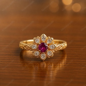 May include: A gold ring with a floral design. The ring features a central round, deep pink gemstone surrounded by white gemstones. The band has an intricate pattern. The ring is displayed on a wooden surface.