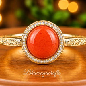 May include: A gold ring featuring a large, round, smooth orange stone at its center, surrounded by a halo of small white stones. The gold band displays an intricate engraved leaf pattern on both sides. The text Bhawanacrafts is visible below.