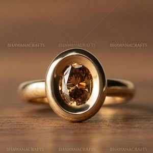 May include: A gold ring featuring a large, oval-cut, brown gemstone set within a bezel. The ring sits on a wooden surface. The band and bezel are gold, creating a cohesive and elegant design. The ring is a piece of jewellery.