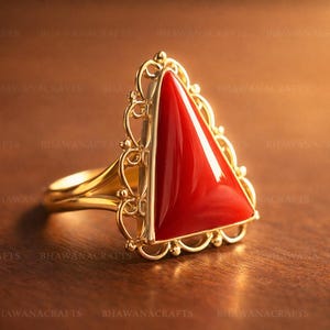 Pode incluir: Um anel de ouro com uma grande pedra preciosa triangular vermelha brilhante. A pedra preciosa está engastada numa moldura de filigrana dourada. A banda do anel também é dourada. O anel é exibido numa superfície de madeira castanha.