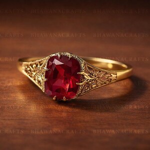 May include: A gold ring featuring a large, rectangular, red gemstone, set within an ornate floral design. The ring is displayed on a dark wooden surface, showcasing the intricate details of the jewellery.