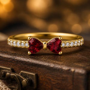 May include: A gold ring featuring a bow-shaped design with two red gemstones and a band encrusted with small, clear gemstones. The ring is set against a blurred, warm-toned background, suggesting a jewelry theme.