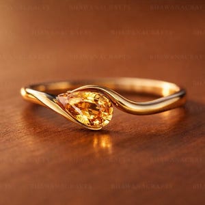 May include: A gold ring with a pear-shaped, yellow gemstone. The ring's band curves gracefully, cradling the stone. The ring is set against a warm, brown background, highlighting the ring's design and color.