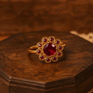May include: A gold ring with a large, round, red gemstone at the center, surrounded by smaller red gemstones in a floral pattern. The ring is displayed on a dark wooden surface.