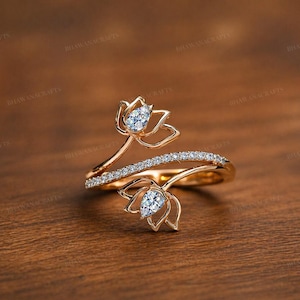 May include: A gold ring with a floral design. Two flower-shaped settings each hold a pear-shaped, clear gemstone. The ring band has a row of small, clear gemstones. The ring is set against a wooden background.