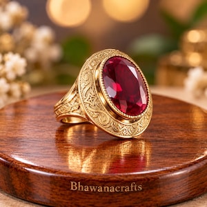 May include: A gold ring featuring a large, oval-cut, deep red gemstone set in a decorative, engraved gold frame. The metal band displays intricate scrollwork patterns surrounding the central stone, which is faceted and reflects light.