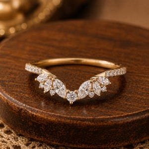 May include: A gold ring with a curved design, set with clear marquise-cut and round gemstones. The band is also set with small, clear gemstones. The ring is displayed on a wooden surface. The ring's design is elegant.