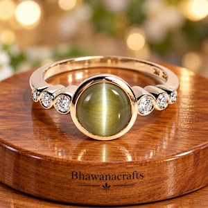 Puede incluir: Un anillo de oro con una piedra preciosa central redonda de ojo de gato verde oliva en talla cabujón con engaste de bisel, flanqueada por tres pequeños diamantes blancos redondos en talla brillante a cada lado, en una banda de oro pulido con el texto Bhawanacrafts.