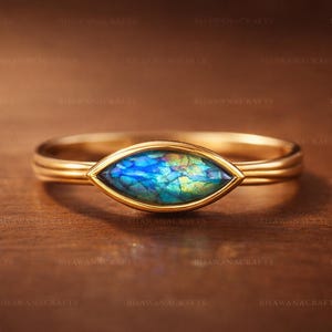 May include: A gold ring with a marquise-shaped, iridescent blue and green stone. The ring band is a simple, thin design. The stone's colors shift with light, creating a unique and eye-catching piece of jewelry.