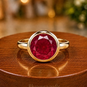 Puede incluir: Un anillo de oro con una gran piedra preciosa de color rojo intenso, redonda y facetada, montada en un engaste de bisel liso. La banda es de oro pulido con un perfil redondeado. El texto Bhawanacrafts es visible en la superficie bajo el anillo.