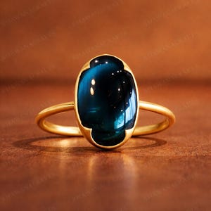 May include: A gold ring featuring an oval, dark teal gemstone. The gemstone is set in a gold bezel with a four-lobed design. The ring's band is a simple, polished gold.