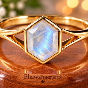 May include: A gold ring featuring a central hexagonal moonstone with a blue iridescent sheen. The band has a split shank design that connects to the bezel setting holding the gemstone. The text Bhawanacrafts is visible on the base below.