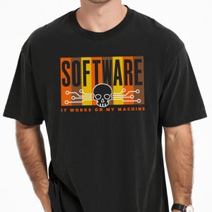 Peut inclure: Un t-shirt noir avec un motif graphique affichant le mot SOFTWARE en lettres noires sur des bandes verticales orange, jaunes et marron. Une icône de crâne blanc avec des lignes de circuit imprimé et le texte IT WORKS ON MY MACHINE.