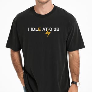 May include: A black short-sleeved t-shirt featuring the white text I IDLE AT 0 dB with a yellow lightning bolt icon replacing the letter E and positioned between the words IDLE and AT. The shirt has a crew neckline and a standard fit.