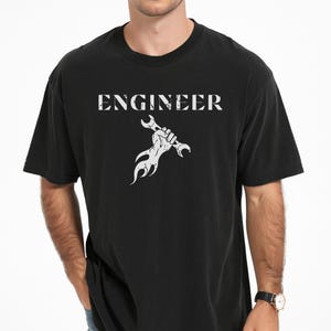 Puede incluir: Una camiseta negra de manga corta con un estampado gráfico blanco en el pecho. El diseño incluye la palabra ENGINEER en una fuente desgastada sobre una mano que sostiene una llave inglesa con llamas saliendo de la parte inferior del puño.