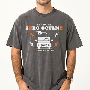 May include: A charcoal gray short-sleeve t-shirt featuring a white and orange graphic of a car with an electric plug icon. The text reads 87 89 93 ZERO OCTANE and ELECTRIC MOTOR CLUB, accompanied by orange and white lightning bolt symbols.