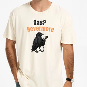 May include: A cream-colored short-sleeve t-shirt featuring a graphic of a black raven holding a microphone. Above the raven, the text reads Gas? in black and Nevermore in orange. The shirt has a crew neckline and a standard fit design.