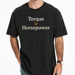 Puede incluir: Una camiseta negra de manga corta con el texto Torque > Horsepower impreso en blanco y naranja en el pecho. La prenda tiene un cuello redondo y un corte estándar, mostrando las palabras en una tipografía serif clara en la parte frontal.