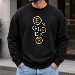 May include: A black long-sleeve crewneck sweatshirt featuring a vertical graphic design. The word ENGINEER is spelled out with the letters E, N, and R enclosed in white gear-shaped icons with yellow centers, interspersed with white letters G, I, E, E.