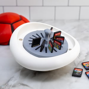 Puede incluir: Un soporte blanco, de forma ovalada, para cartuchos de juegos con un interior gris, que contiene varios cartuchos de juegos. Un balón de fútbol rojo está en el fondo. También se ven varios cartuchos de juegos sueltos.