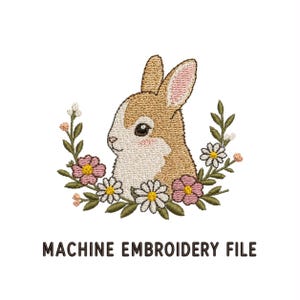 May include: A machine embroidery file featuring a tan and white rabbit head surrounded by a semi-circular wreath of pink flowers, white daisies, and green leaves. The text "MACHINE EMBROIDERY FILE" is printed in black capital letters below the design.