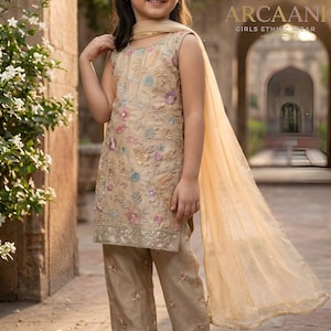 May include: A young girl wearing a beige ethnic wear outfit with floral embroidery. The outfit includes a sleeveless top, matching pants, and a sheer scarf. The outfit is from the brand ARCAANI, Girls Ethnic Wear.