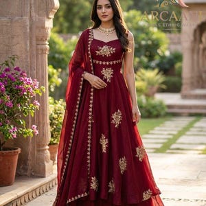 Puede incluir: Un vestido étnico burdeos con bordados dorados. El vestido tiene un escote en forma de corazón y una falda fluida. Una bufanda a juego está drapeada sobre el hombro. La marca "ARCAANI GIRLS ETHNIC WEAR" es visible.