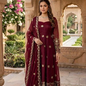 May include: A deep maroon floor-length dress with long sleeves, featuring gold floral embroidery across the bodice, sleeves, and hem. A matching sheer maroon dupatta with a gold embroidered border is draped over one shoulder. Text reads ARCAANI.