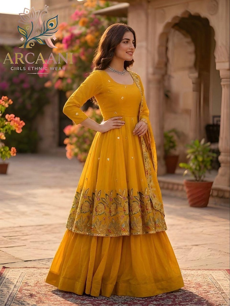 Puede incluir: Un vestido &eacute;tnico amarillo vibrante con un dobladillo bordado floral. El vestido presenta una parte superior de manga larga y una falda en capas. La marca "ARCAANI GIRLS ETHNIC WEAR" es visible.