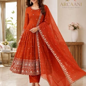 May include: A vibrant orange long-sleeved dress with a flared skirt, featuring intricate silver embroidery along the hem and neckline. The outfit includes matching orange trousers and a sheer, embroidered orange dupatta draped over the shoulder.