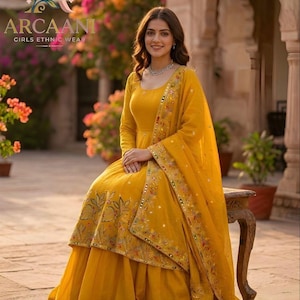 Puede incluir: Una mujer con un atuendo &eacute;tnico amarillo mostaza. El conjunto incluye un vestido de manga larga y una dupatta a juego, ambos adornados con intrincados bordados. El vestido presenta una falda acampanada y se combina con un delicado collar. La marca "ARCAANI" es visible.