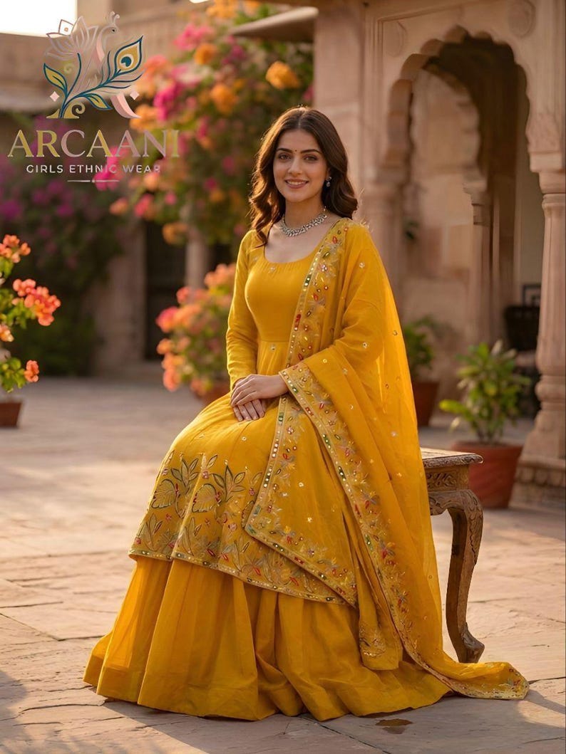 Puede incluir: Una mujer con un atuendo &eacute;tnico amarillo mostaza. El conjunto incluye un vestido de manga larga y una dupatta a juego, ambos adornados con intrincados bordados. El vestido presenta una falda acampanada y se combina con un delicado collar. La marca "ARCAANI" es visible.
