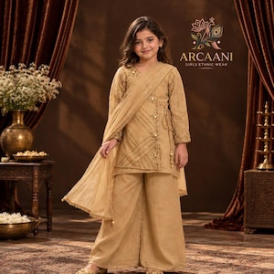 Könnte beinhalten: Eine beige langärmelige Tunika mit diagonaler Stickerei und seitlichen Bindebändern, kombiniert mit einer passenden weiten Hose und einem transparenten Dupatta. Das Kleidungsstück trägt das Logo Arcaani Girls Ethnic Wear.