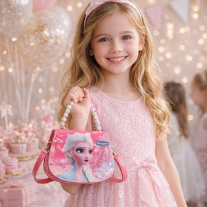 May include: A young girl in a pink dress holds a pink purse featuring a Frozen movie design. The purse has a beaded handle and a shoulder strap. The girl is smiling and wearing a matching pink headband. The background includes party decorations.