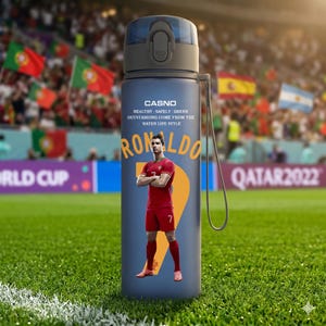 May include: A blue water bottle featuring a portrait of Cristiano Ronaldo in a red soccer uniform. The bottle has a black cap and a strap. Text on the bottle includes "CASNO" and "RONALDO". The background shows a soccer field and flags.