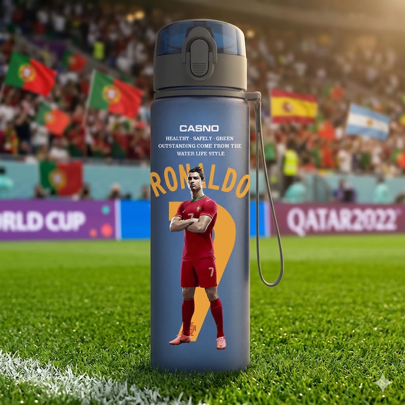 May include: A blue water bottle featuring a portrait of Cristiano Ronaldo in a red soccer uniform. The bottle has a black cap and a strap. Text on the bottle includes "CASNO" and "RONALDO". The background shows a soccer field and flags.