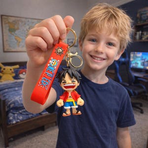 Puede incluir: Un llavero rojo con el texto "ONE PIECE" en blanco y un llavero dorado. El llavero presenta una pequeña figura de Luffy con una chaqueta roja, pantalones cortos azules y zapatos negros. El fondo incluye un mapa y un niño.