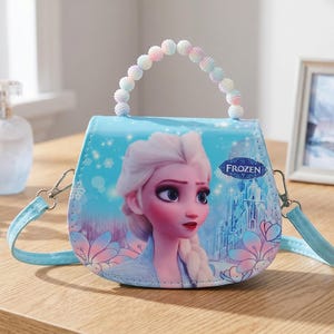 May include: A light blue purse featuring a graphic of Elsa from the movie Frozen. The purse has a beaded handle and a detachable shoulder strap. The word "FROZEN" is printed on the front. Other items on the wooden surface include perfume bottles and a framed picture.