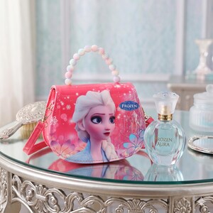 May include: A pink purse featuring the Frozen character Elsa, with a beaded handle and a shoulder strap. Next to it is a clear perfume bottle with a gold cap and the text "Rozen Aura". The items are on a reflective surface.