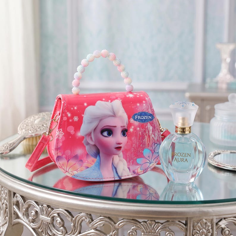 May include: A pink purse featuring the Frozen character Elsa, with a beaded handle and a shoulder strap. Next to it is a clear perfume bottle with a gold cap and the text "Rozen Aura". The items are on a reflective surface.