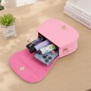 May include: A pink, small, open purse with a gold clasp, filled with items like a phone, glasses, and cosmetics. The purse is on a wooden surface. A small succulent plant in a glass container is visible in the background.